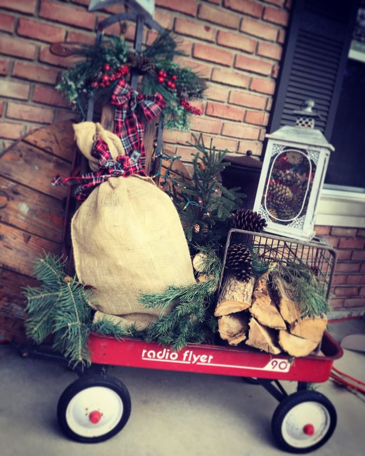 a red wagon filled with logs and pine cones next to a brick wall covered in christmas decorations