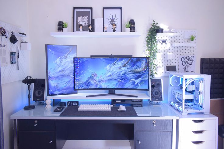 a desk with two computer monitors and speakers on it, in front of a white wall