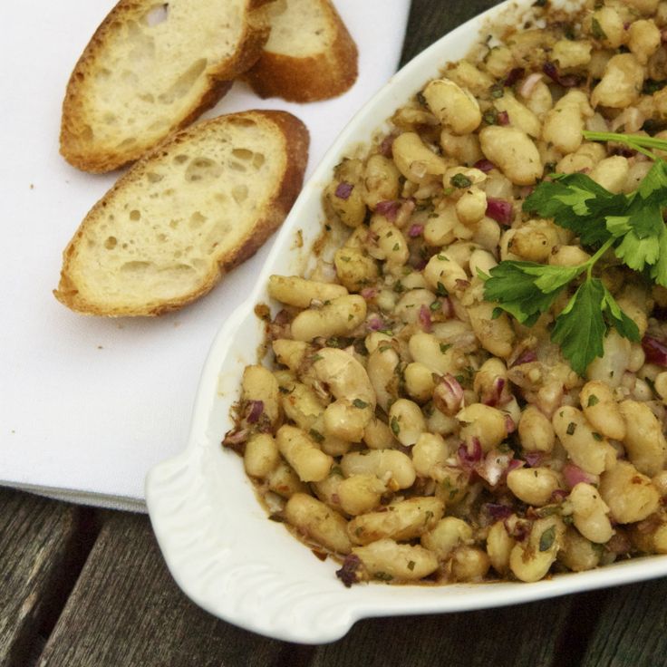 a white dish filled with beans and garnished with parsley next to two slices of bread