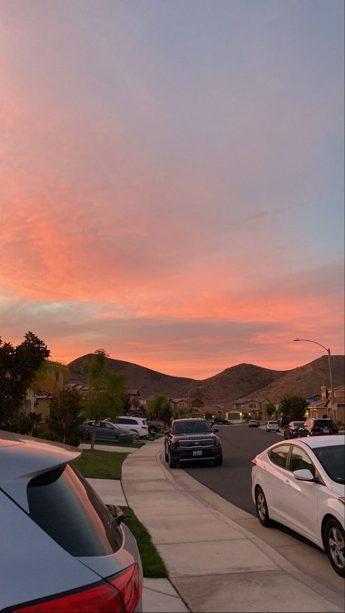 cars parked on the side of a road at sunset with mountains in the background and pink clouds