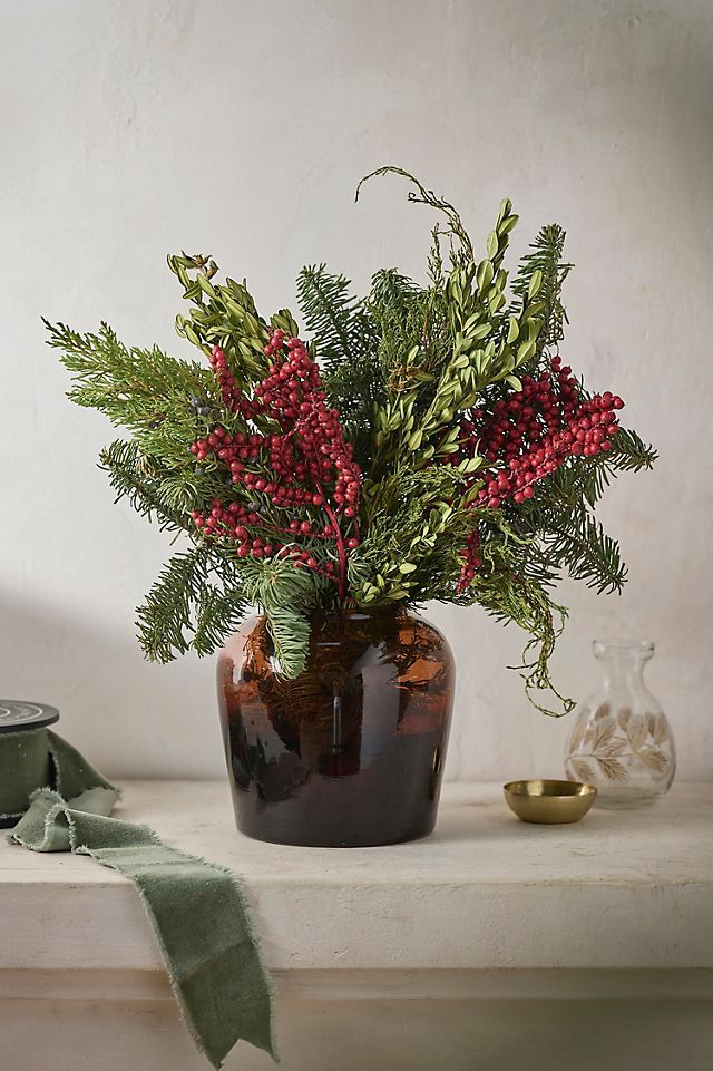 a vase filled with red berries and greenery on top of a white shelf next to a candle