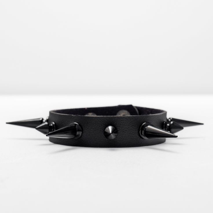 This bracelet measures 9 inches long and 1 inch wide, and is crafted from black, supple leather for maximum comfort. The adjustable metal button buckle allows for a customizable fit. Adorned with 4 large, glossy black alloy metal studs that protrude 1 inch, as well as 3 smaller studs made from the same luxurious material. Edgy Black Metal Cuff Bracelet, Black Punk Bracelets, Edgy Black Bracelet Strap Jewelry, Punk Black Bracelets, Punk Black Bracelet, Black Edgy Jewelry With Bracelet Strap, Edgy Black Jewelry With Bracelet Strap, Edgy Black Metal Wristband, Punk Leather Jewelry With Black Band