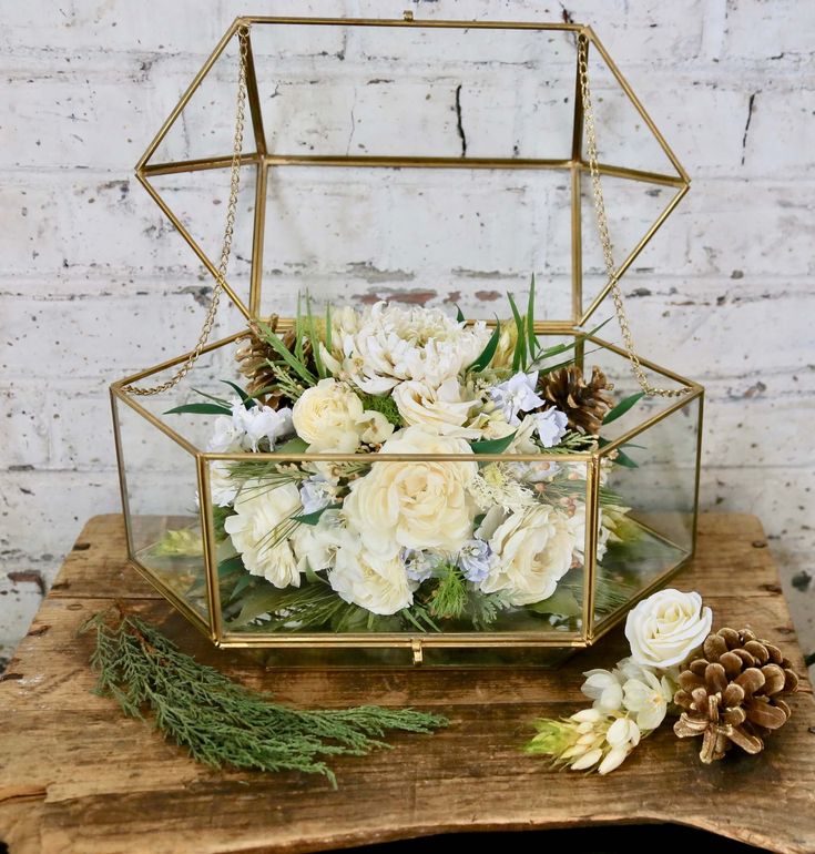 a glass box filled with white flowers and pine cones on top of a wooden table