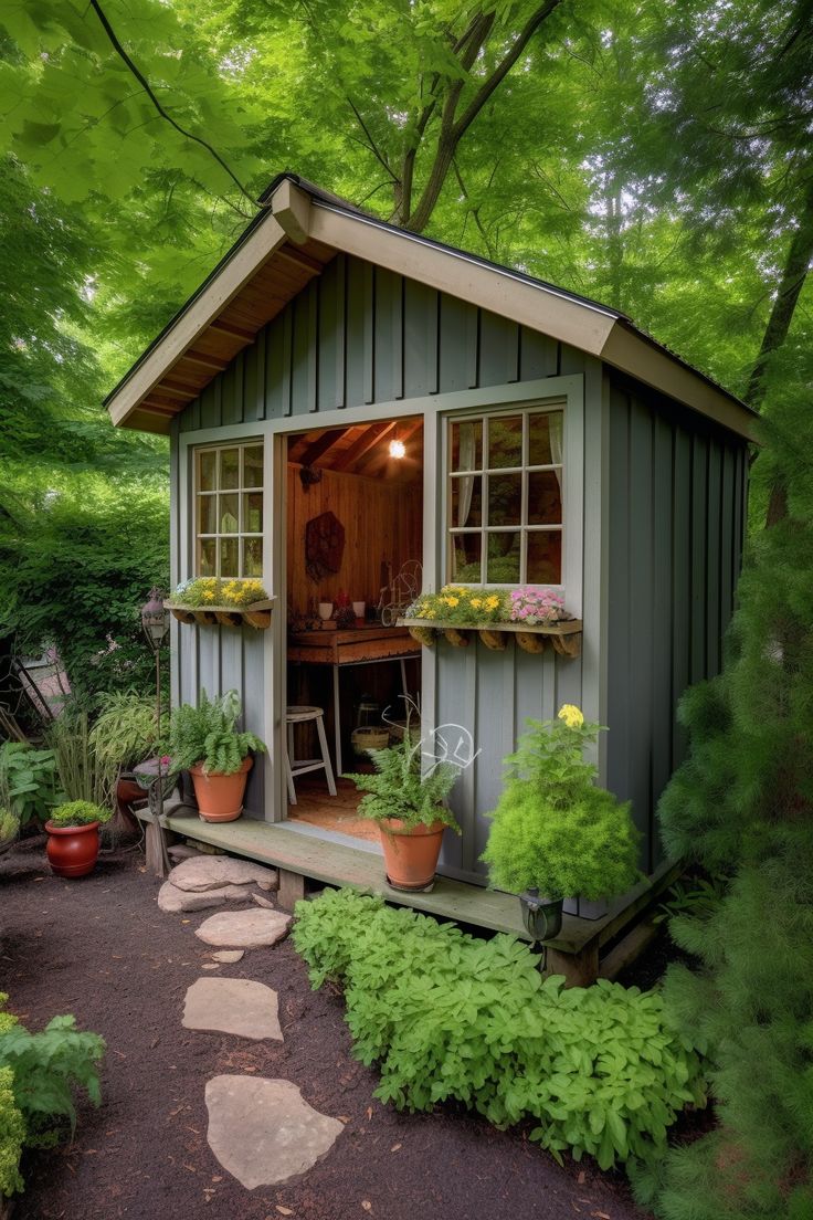 a small shed with potted plants in the front and windows on the side, surrounded by greenery