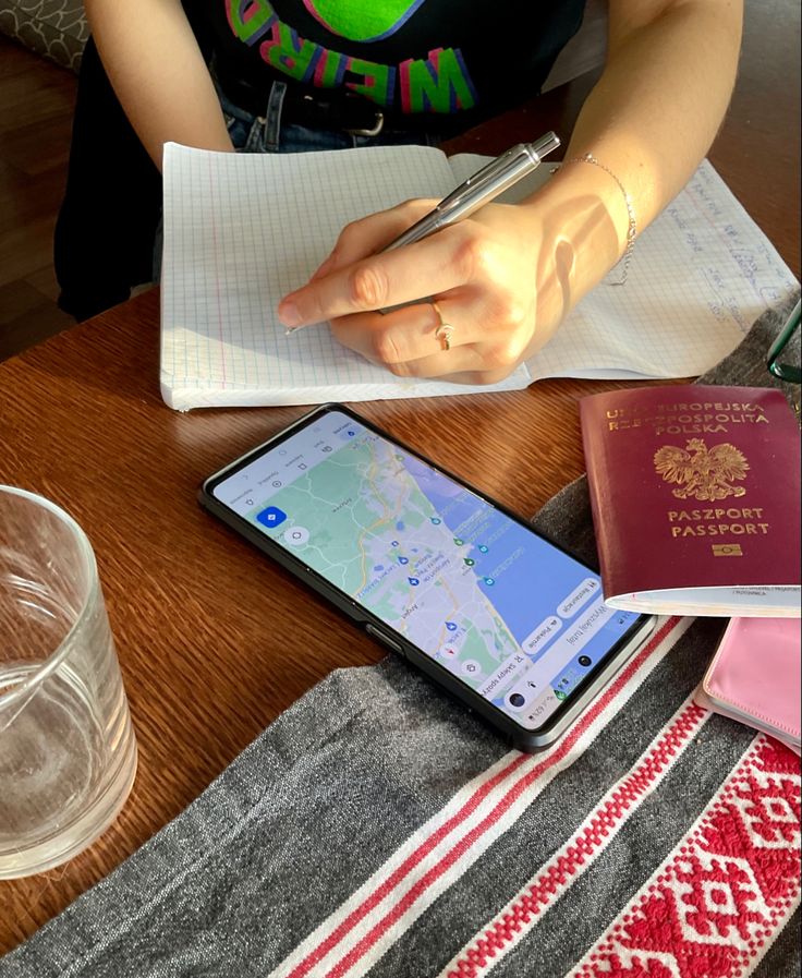 a person sitting at a table with a cell phone and passport, drinking water, and holding a pen