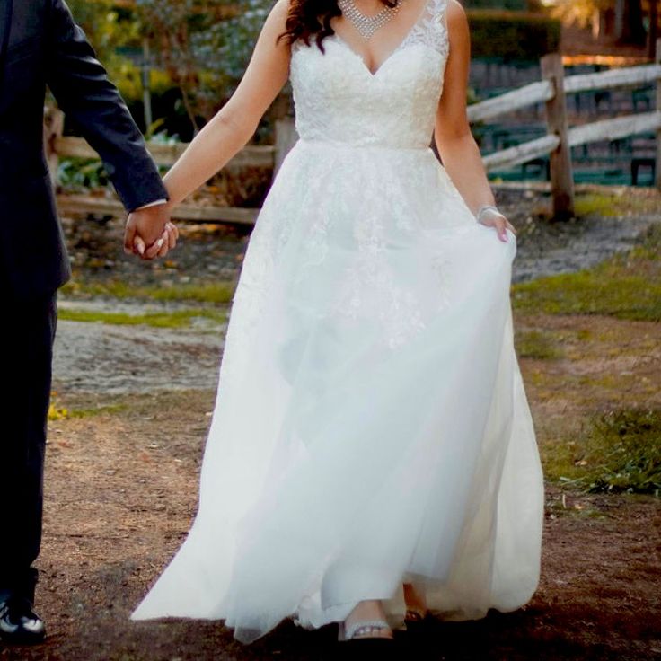 a bride and groom hold hands as they walk down a path in their wedding attire