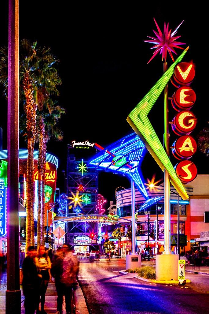 people are walking down the street in front of neon signs and palm trees at night