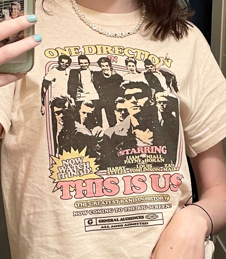 a woman holding a cell phone in her right hand and wearing a t - shirt with the words one direction on it