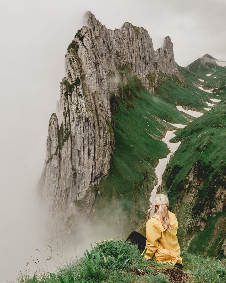 a woman sitting on top of a lush green hillside next to a tall rock formation