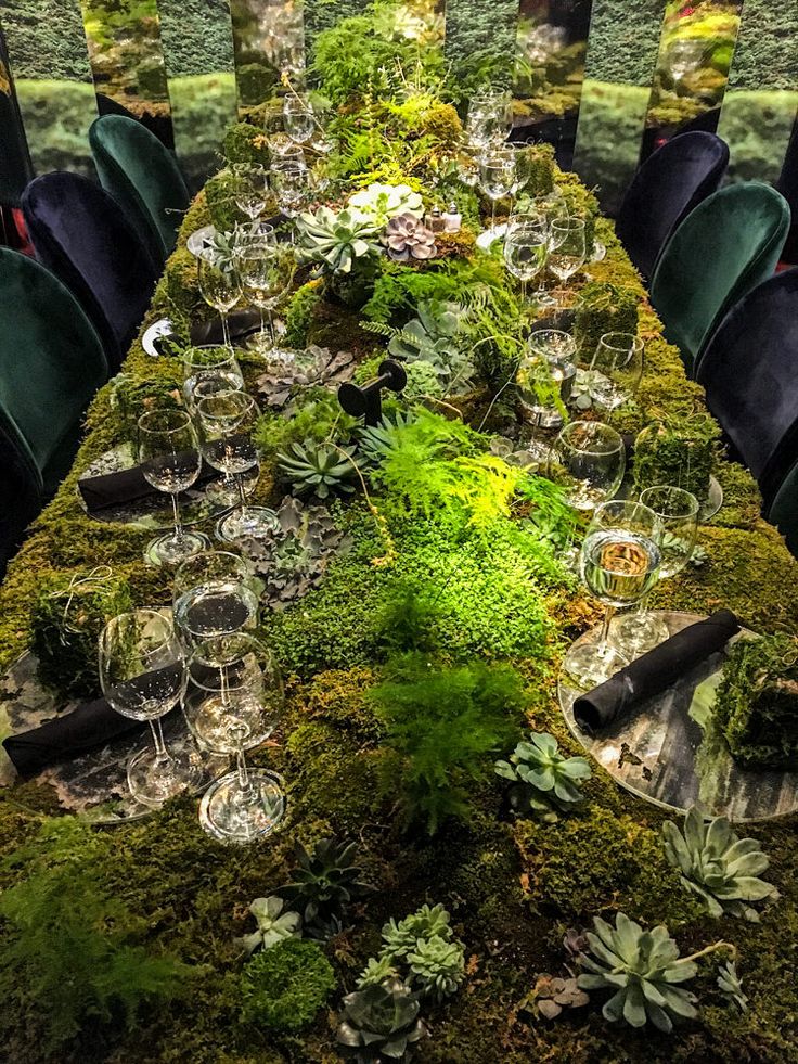 a long moss covered table with wine glasses and plants in the center is surrounded by green chairs