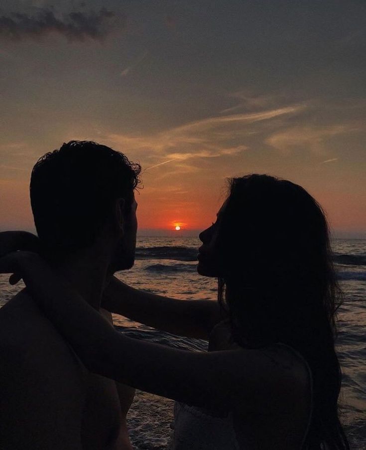 a man and woman are standing on the beach as the sun sets in the background