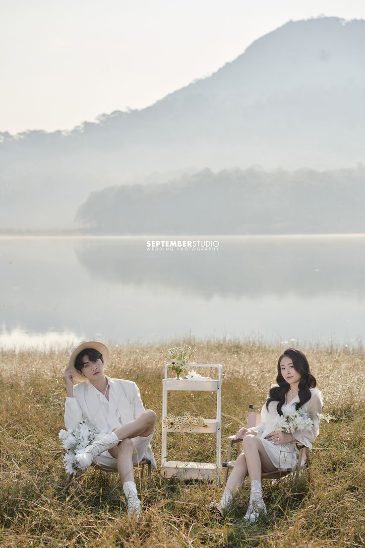 two women sitting in the grass next to each other near a body of water with mountains in the background