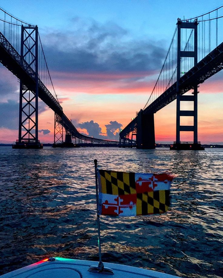 a flag on the back of a boat in front of a suspension bridge at sunset