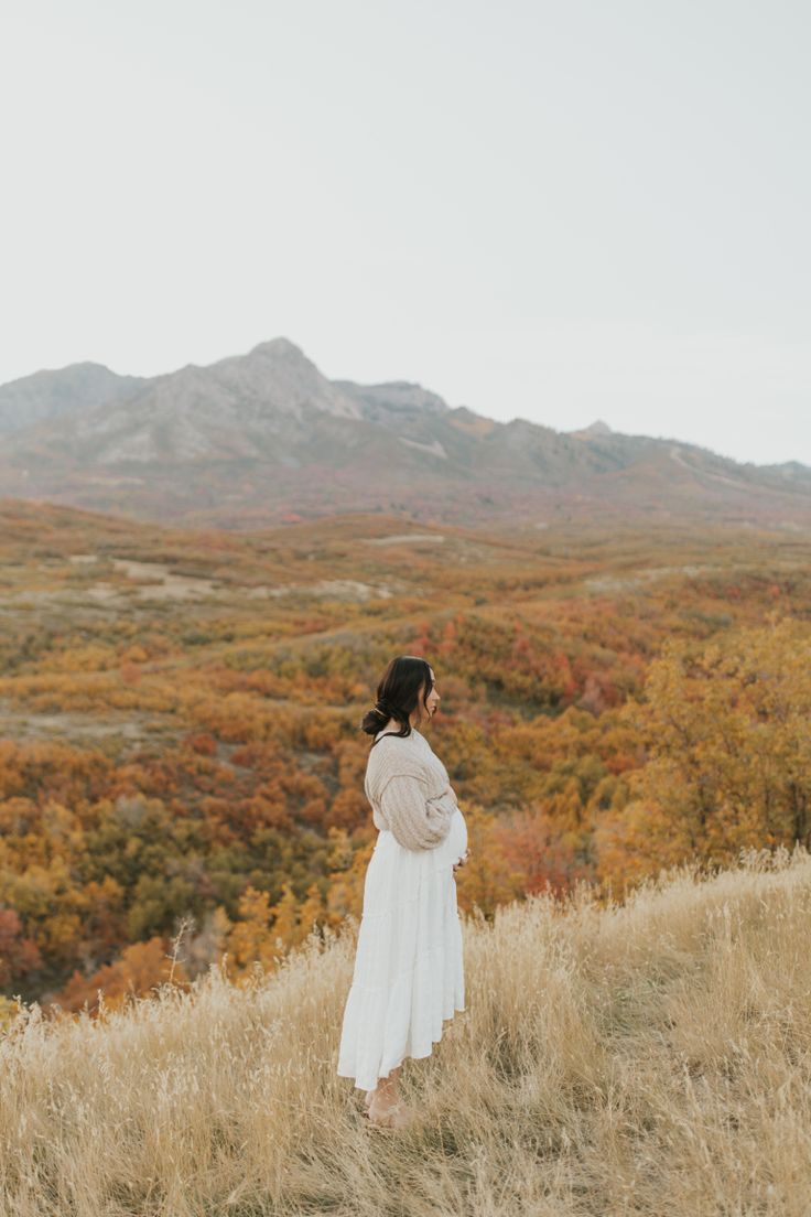 a woman standing on top of a grass covered hillside in front of a mountain range