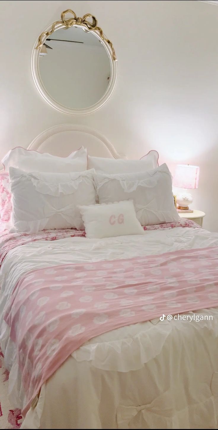 a white bed with pink and white comforter in a bedroom next to a round mirror