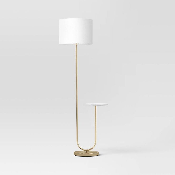 a lamp with a white shade on it and a gold stand in front of the light