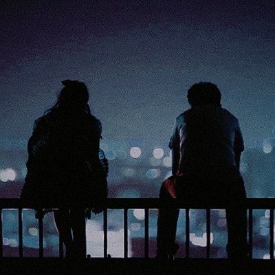 two people standing next to each other on a balcony at night with city lights in the background