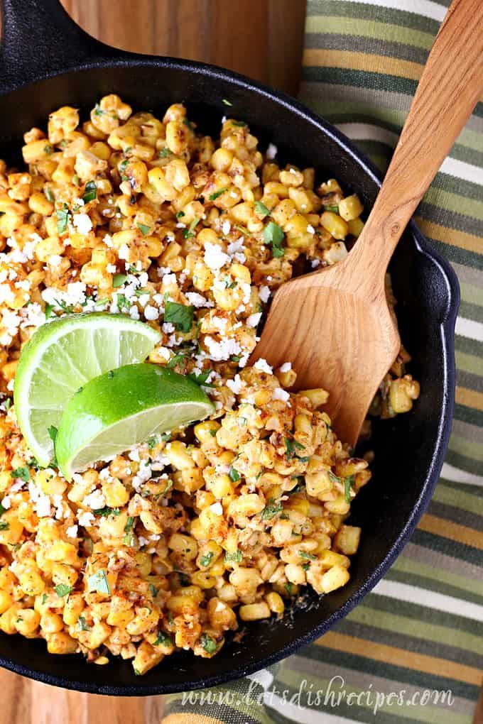 mexican street corn in a cast iron skillet with a wooden spoon and lime wedge