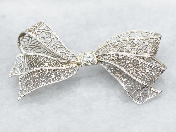 Add a touch of elegance and whimsy to any outfit with this bow brooch. The delicate filigree detailing and sparkling European Cut diamond make this brooch a timeless statement piece. Perfect for any occasion!Metal: 14K White GoldGem: European Cut Diamond .10 Carats, SI1 in Clarity, I in ColorGem Measurements: 3.2 mm, RoundMeasurements: 44 x 23 mmMarks: "X14K" Stamped on the pin guard Elegant White Gold Brooch With Intricate Design, Elegant White Gold Brooches With Intricate Design, Elegant Wedding Jewelry With Decorative Bow, Diamond Filigree Brooches As Gift, Elegant Filigree Brooch For Anniversary, Ornate White Gold Brooches For Wedding, Elegant Filigree Brooches For Anniversary, Elegant Filigree Brooches For Evening, Ornate White Gold Wedding Brooches