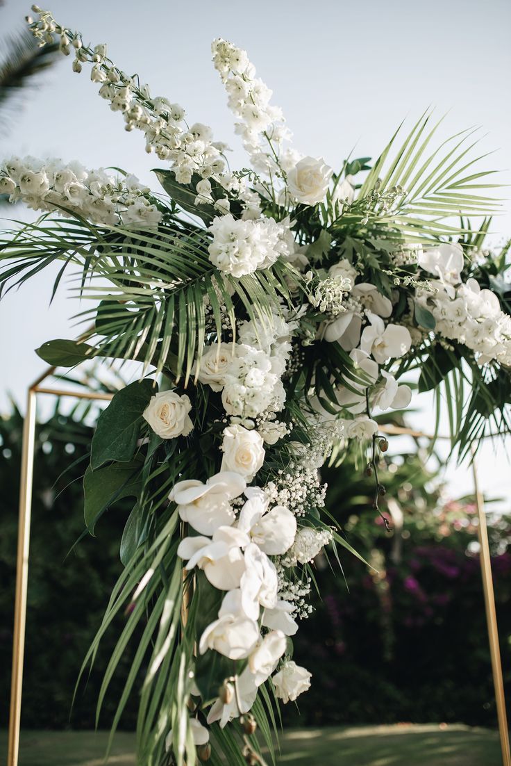 white flowers and greenery are arranged on a gold frame for an outdoor wedding ceremony