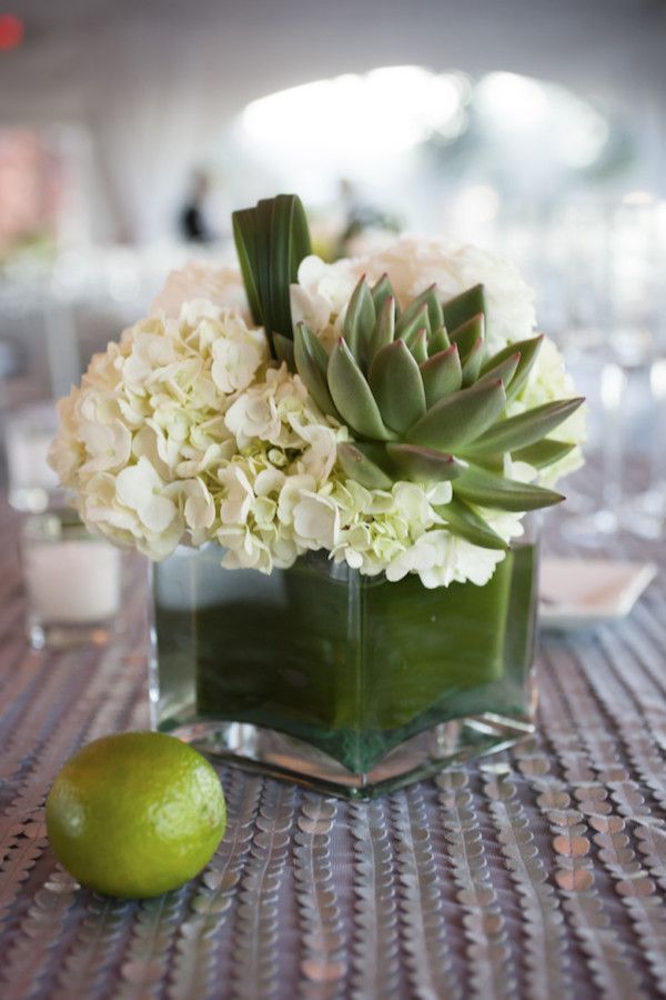 an arrangement of white flowers and green leaves in a square glass vase on a table