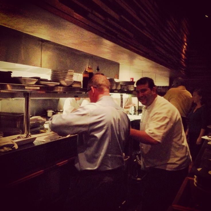 two men in a kitchen preparing food for people to eat at the counter and behind them is another man