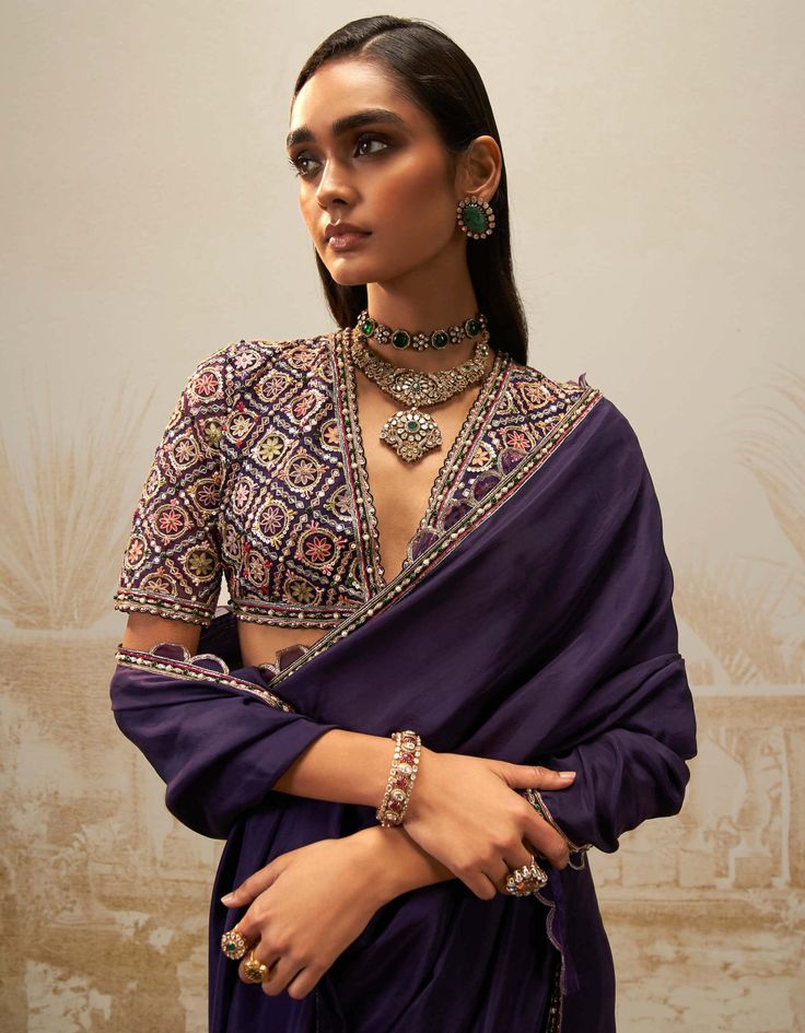 Elevate your ethnic wardrobe with this purple geometric patterns sari set – a seamless blend of tradition and trend. This ensemble features a silk flat sari, petticoat, and a purple v neck embroidered blouse adorned with vibrant multicolor threadwork. The regal purple hue is versatile for any occasion, while geometric patterns and scalloped detailing bring a modern flair. Effortlessly elevate your style with this chic sari set. Dark Purple Saree Blouse Combination, Light Purple Saree Blouse Combination, Purple Saree Sets With Traditional Patterns, Bohemian Purple Saree With Traditional Patterns, Purple Sari With Contrast Blouse, V Neck Blouse Indian, Purple Saree With Motifs, V Neck Blouse Designs, Purple Embroidered Saree