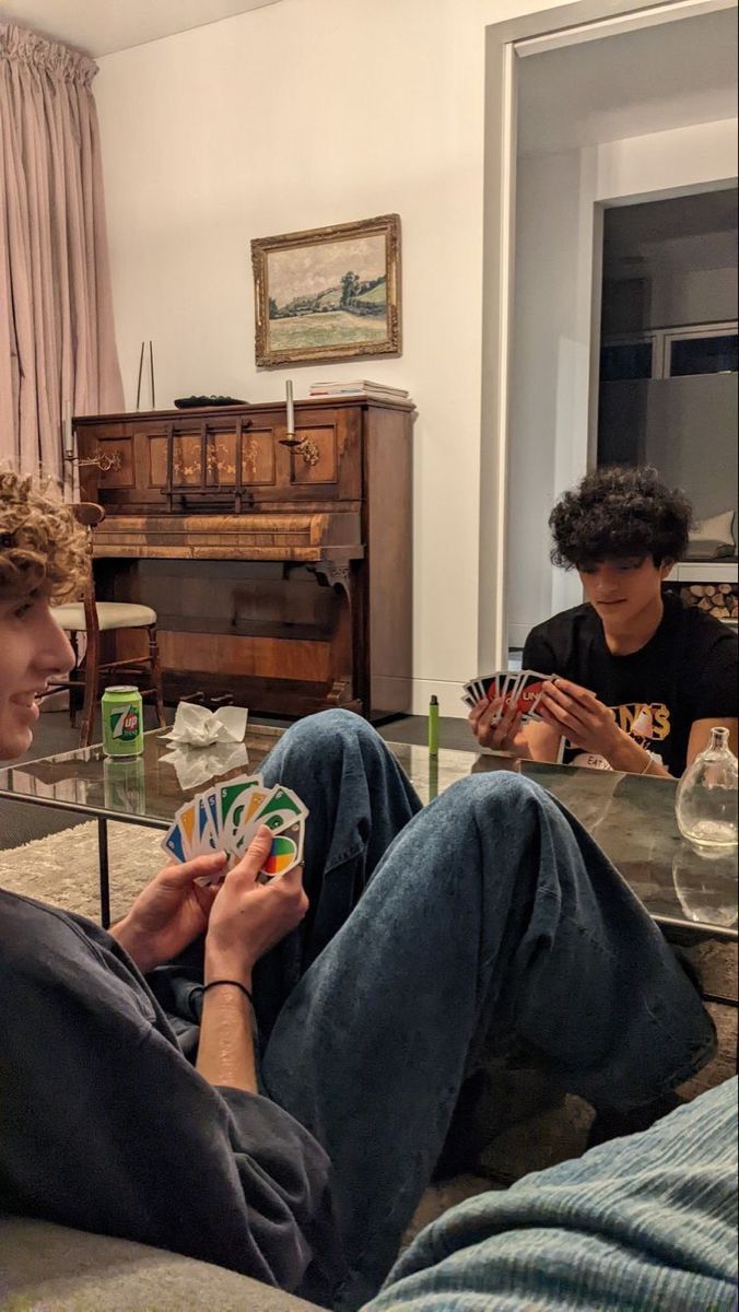 two young men sitting on a couch playing cards in front of a glass coffee table