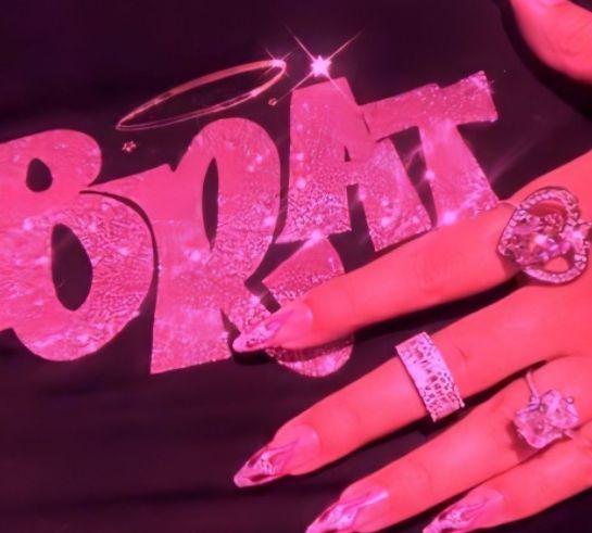 a woman with pink nails and rings on her hand, wearing a black shirt that says brat