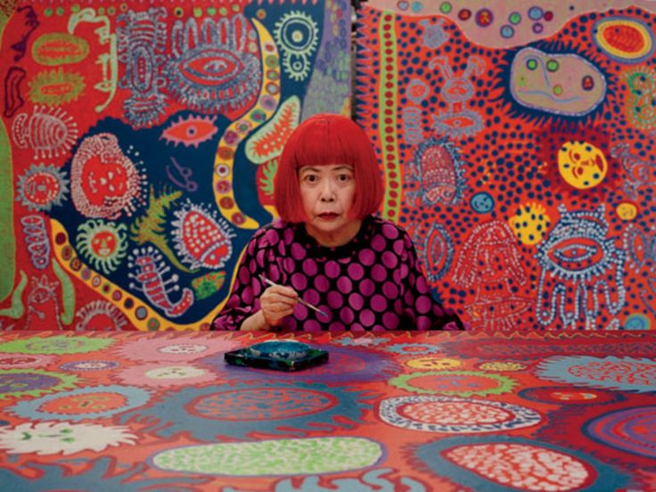 a woman with red hair sitting at a table covered in art