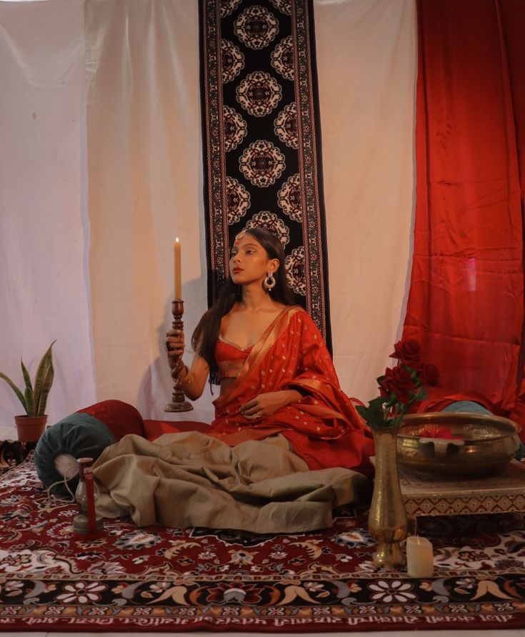 a woman sitting on top of a rug holding a candle in her hand and wearing a red sari