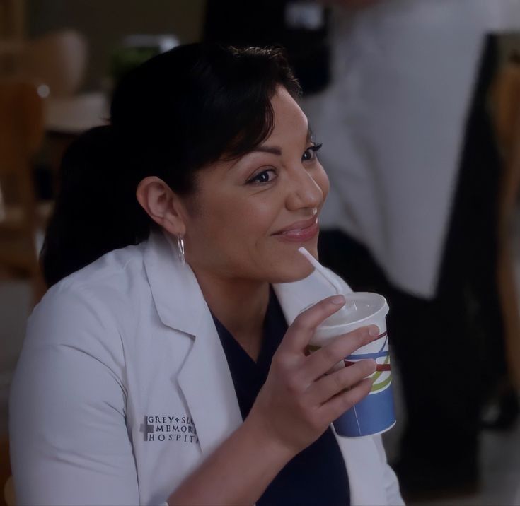 a woman in a white coat is drinking from a cup and smiling at the camera