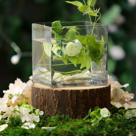 a glass vase filled with flowers on top of a wooden stump in the grass and surrounded by greenery