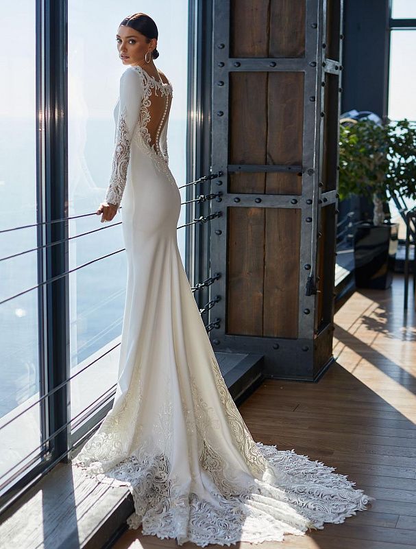 a woman standing in front of a window wearing a long sleeved wedding dress with open back