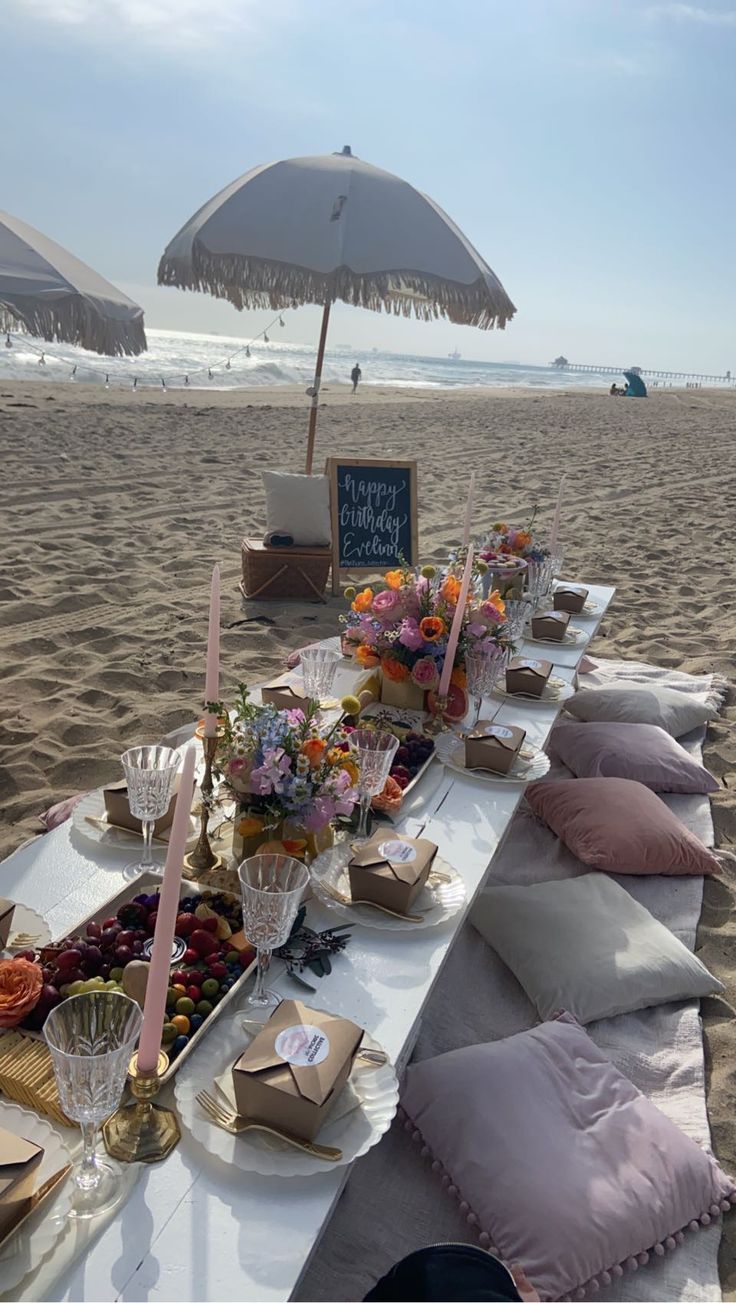 a table set up on the beach with an umbrella over it and other food items