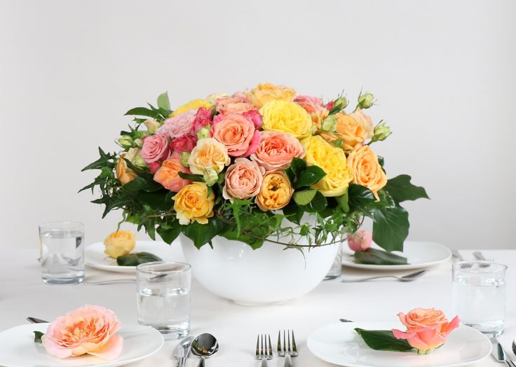 an arrangement of flowers in a white bowl on a table with place settings and silverware