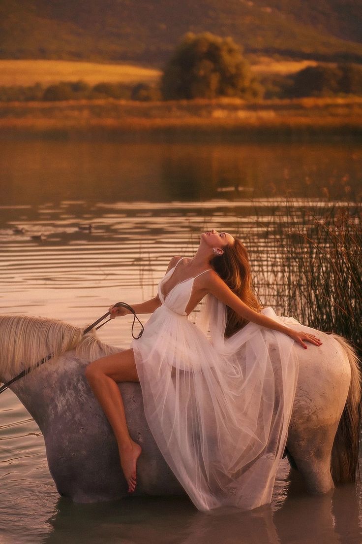 a woman in white dress riding on the back of a horse through water at sunset