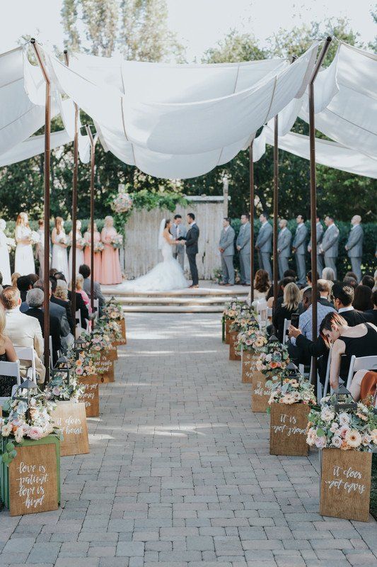 an outdoor ceremony with white draping and flowers on the aisle, surrounded by people sitting in chairs