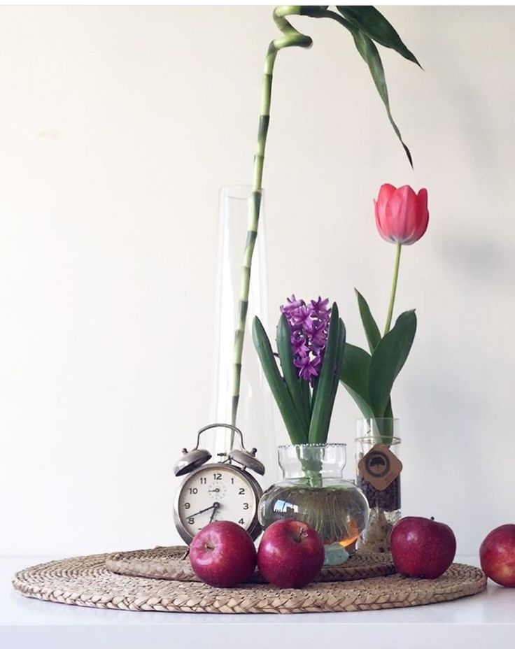 an arrangement of flowers and apples on a table with a clock in the middle,