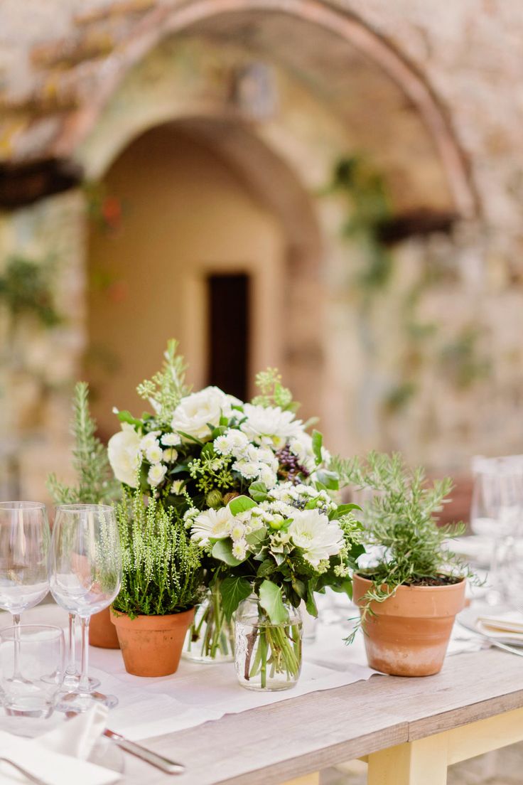 the table is set with white flowers and greenery in terracotta vases