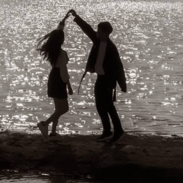 a man and woman standing next to each other near the water's edge with their arms in the air