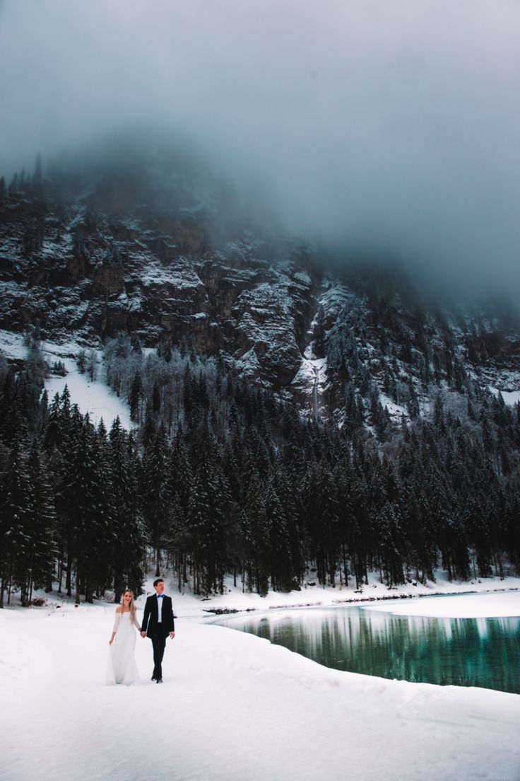 a bride and groom are walking in the snow near a mountain lake with pine trees