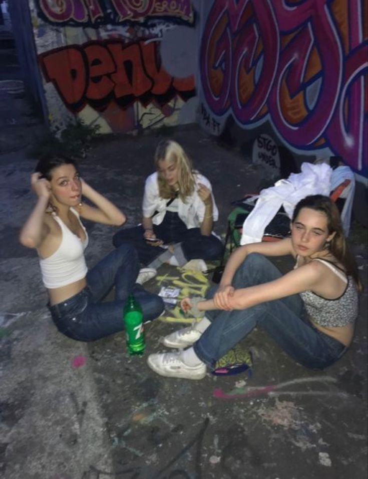 three young women sitting on the ground in front of graffiti covered walls, one is holding her head