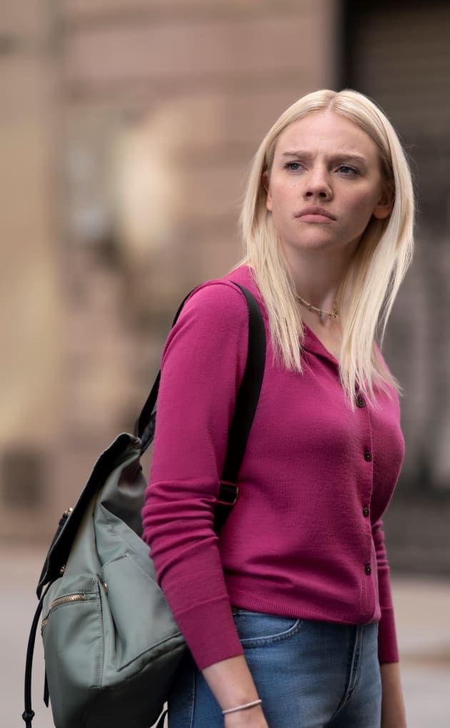 a woman with blonde hair is holding a backpack and looking off to the side while standing in front of a building