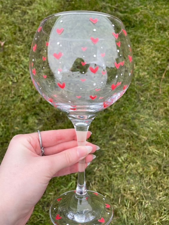 a hand holding a wine glass with hearts painted on the outside and inside, in front of grass