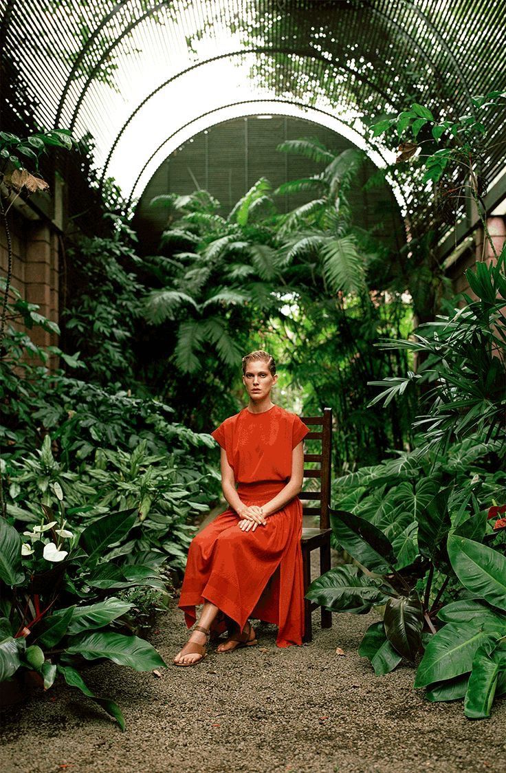 a woman in an orange dress is sitting on a bench surrounded by plants and greenery