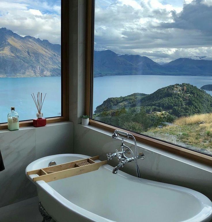 a bath tub sitting under a window next to a large body of water with mountains in the background