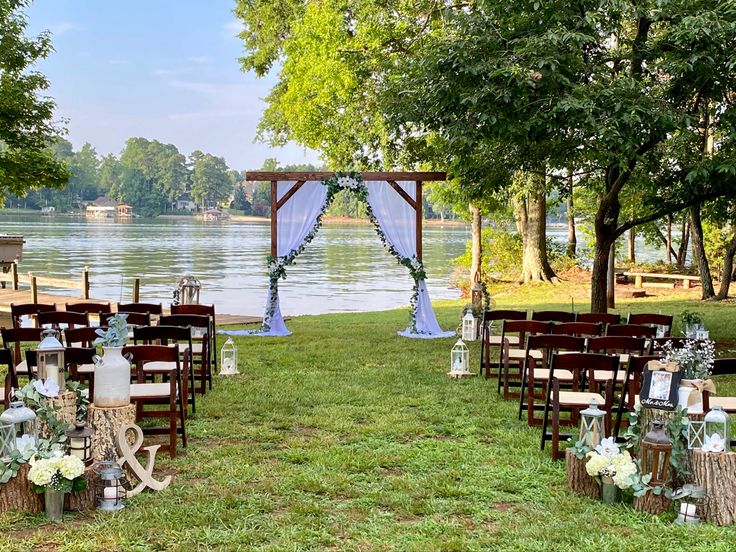 an outdoor ceremony set up with chairs and flowers on the grass next to water in front of trees