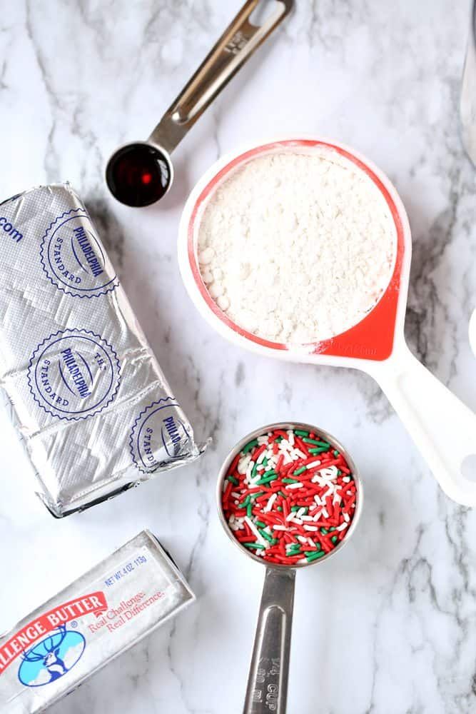 ingredients to make christmas cookies sitting on a marble counter with measuring spoons and flour