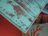 an old wooden table with red and blue paint on it's top, next to a basket filled with pillows